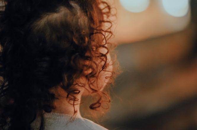 Foto horizontal: detalhe em plano fechado visto de costas e de perfil de criança com cabelo escuro e encaracolado iluminado pela luz dourada, com fundo desfocado.