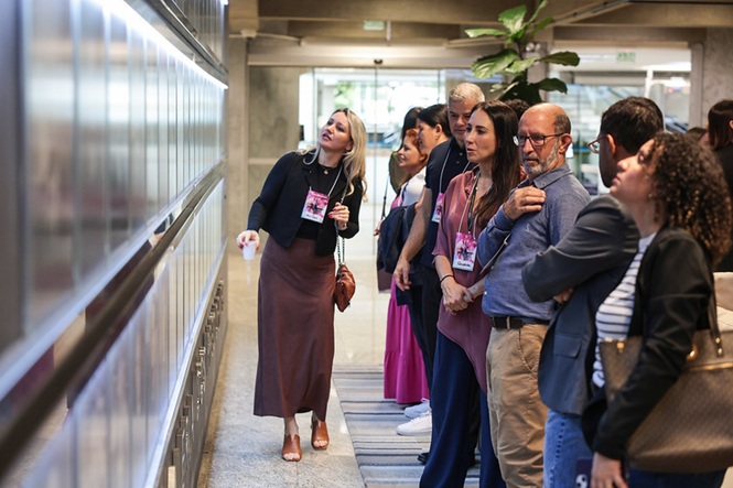 Foto horizontal: grupo de visitantes e servidores com crachás em trajes formais e casuais nas cores preto, marrom, azul e bege observa atentamente galeria iluminada em parede metálica no corredor interno de edifício institucional com piso claro e planta ao fundo durante visita guiada.