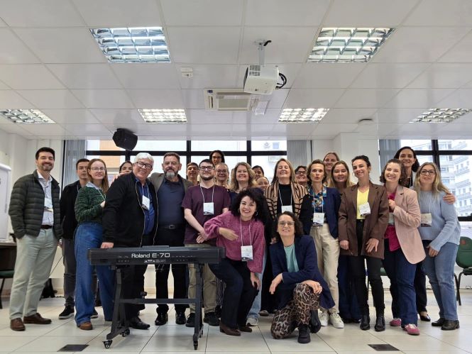 Foto de um grupo de pessoas reunidas em uma sala de aula, posando para a câmera. Ao centro, há um teclado eletrônico e duas pessoas posicionadas à frente do grupo. Ao fundo, aparecem janelas, mesas, cadeiras, luminárias e um projetor instalado no teto.