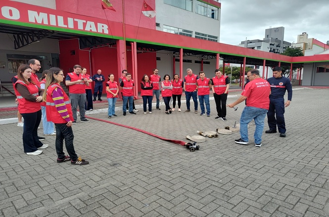 Foto: grupo em coletes vermelhos assiste a bombeiro com mangueiras no pátio do batalhão em treinamento de brigada.