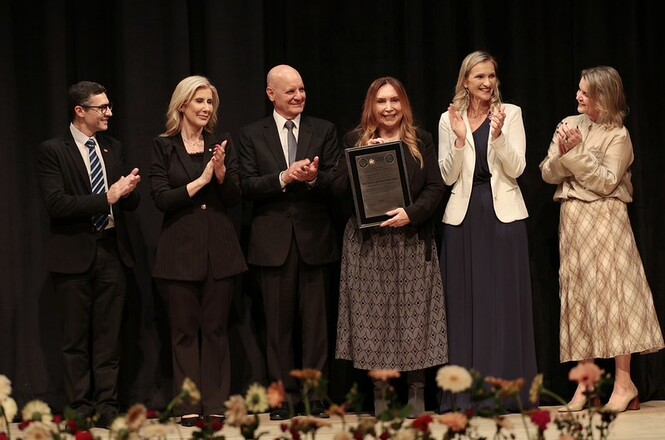 Foto horizontal: mulher em casaco preto e saia estampada segura placa de homenagem e posa ao lado de autoridades em trajes formais pretos, branco e bege em palco com fundo preto e flores em primeiro plano durante cerimônia solene de premiação.