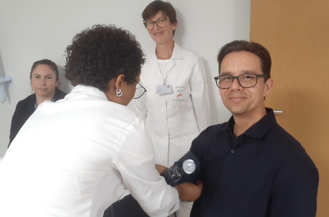 Foto horizontal: homem em camisa preta tem a pressão arterial aferida por profissional de saúde de jaleco branco em consultório médico.
