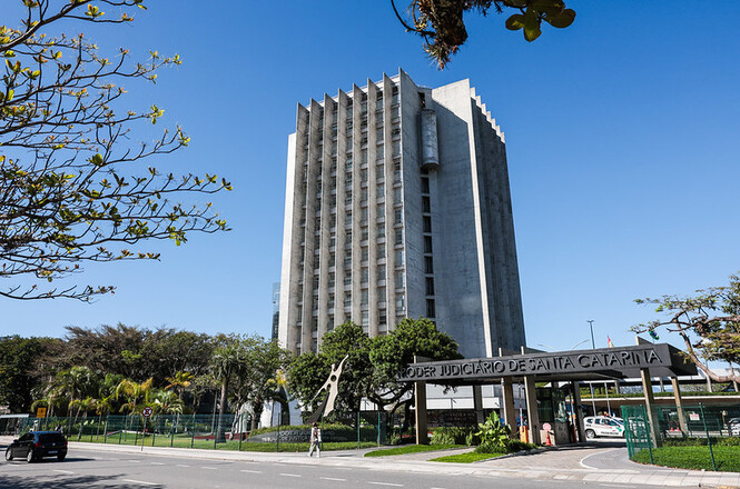 Foto horizontal: prédio cinza sob céu azul e árvores verdes exibe a sede do tribunal em dia ensolarado.
