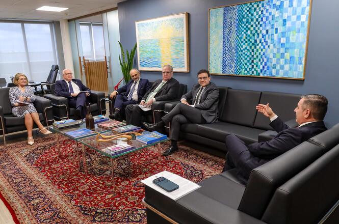 Foto horizontal: autoridades em ternos escuros reúnem-se em sala com sofás pretos e tapete vermelho em reunião oficial.