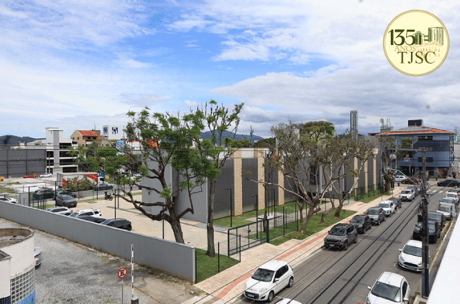 Foto horizontal: prédio cinza sob céu azul em plano aéreo mostra rua movimentada com carros estacionados.