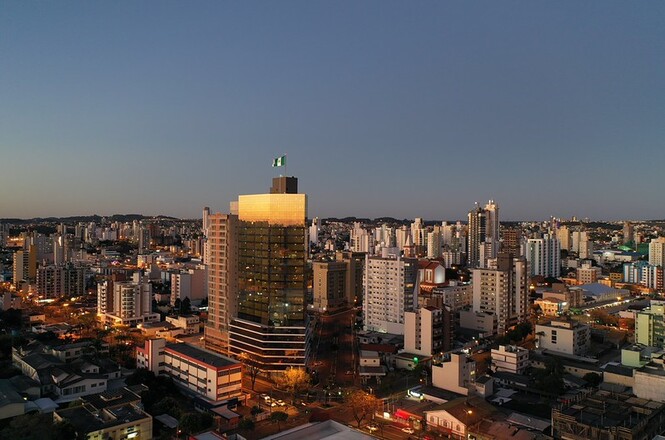 Foto horizontal: prédios sob céu azul e luz dourada em plano aéreo destacam a paisagem urbana ao entardecer.