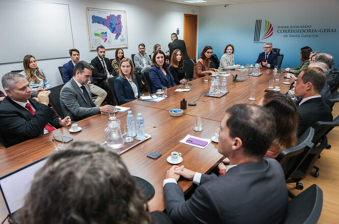 Foto horizontal: grupo numeroso de magistrados e servidores em trajes formais nas cores preto, azul-marinho, cinza, marrom e branco senta-se ao redor de longa mesa oval de madeira em sala de reuniões iluminada, com mapa de Santa Catarina e logotipo da Corregedoria-Geral da Justiça na parede azul ao fundo durante reunião institucional