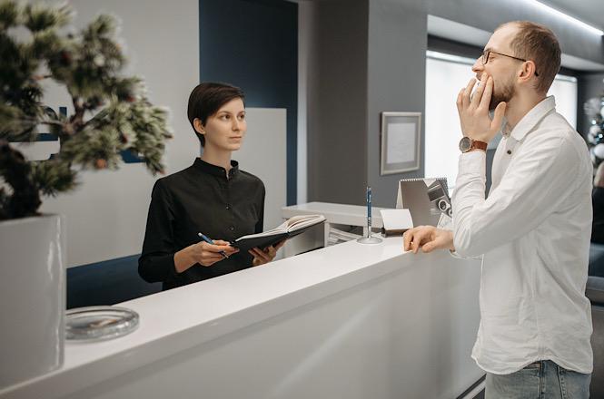 Foto horizontal: homem em camisa branca e óculos leva a mão ao rosto com expressão de dor ao relatar incômodo para recepcionista em camisa preta segurando prancheta e caneta, separados por balcão de atendimento branco em recepção de clínica