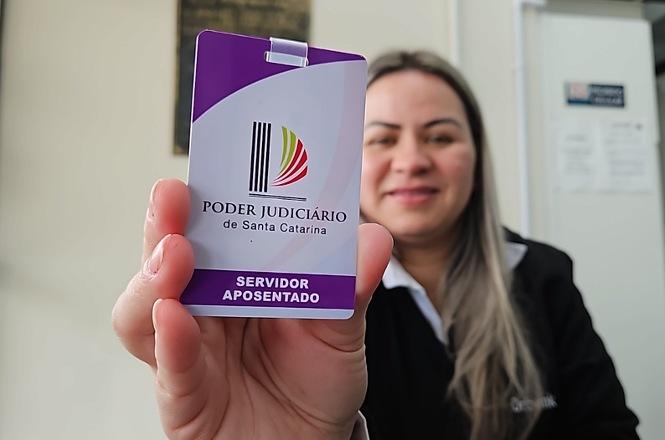 Foto horizontal: mulher sorridente em vestimenta preta com gola branca exibe crachá funcional em primeiro plano com detalhes em roxo, branco, preto, verde e vermelho, contendo o logotipo e os dizeres "Poder Judiciário de Santa Catarina - Servidor Aposentado".
