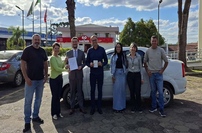 Foto horizontal: grupo em trajes sociais e casuais nas cores cinza, preto, verde-claro, azul e branco posa alinhado em pátio pavimentado diante de automóvel branco, com árvores, céu azul e estabelecimento comercial ao fundo durante ato de entrega de veículo, exibindo documentos oficiais.