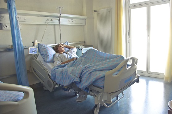 Mulher deitada em cama de hospital coberta por uma manta azul olhando para o lado de fora com a mão esquerda na cabeça