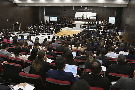 Homens e mulheres sentados em cadeiras almofadadas da cor bordô no Pleno do Tribunal de Justiça do Estado de Santa Catarina olhando para a mesa de autoridades.